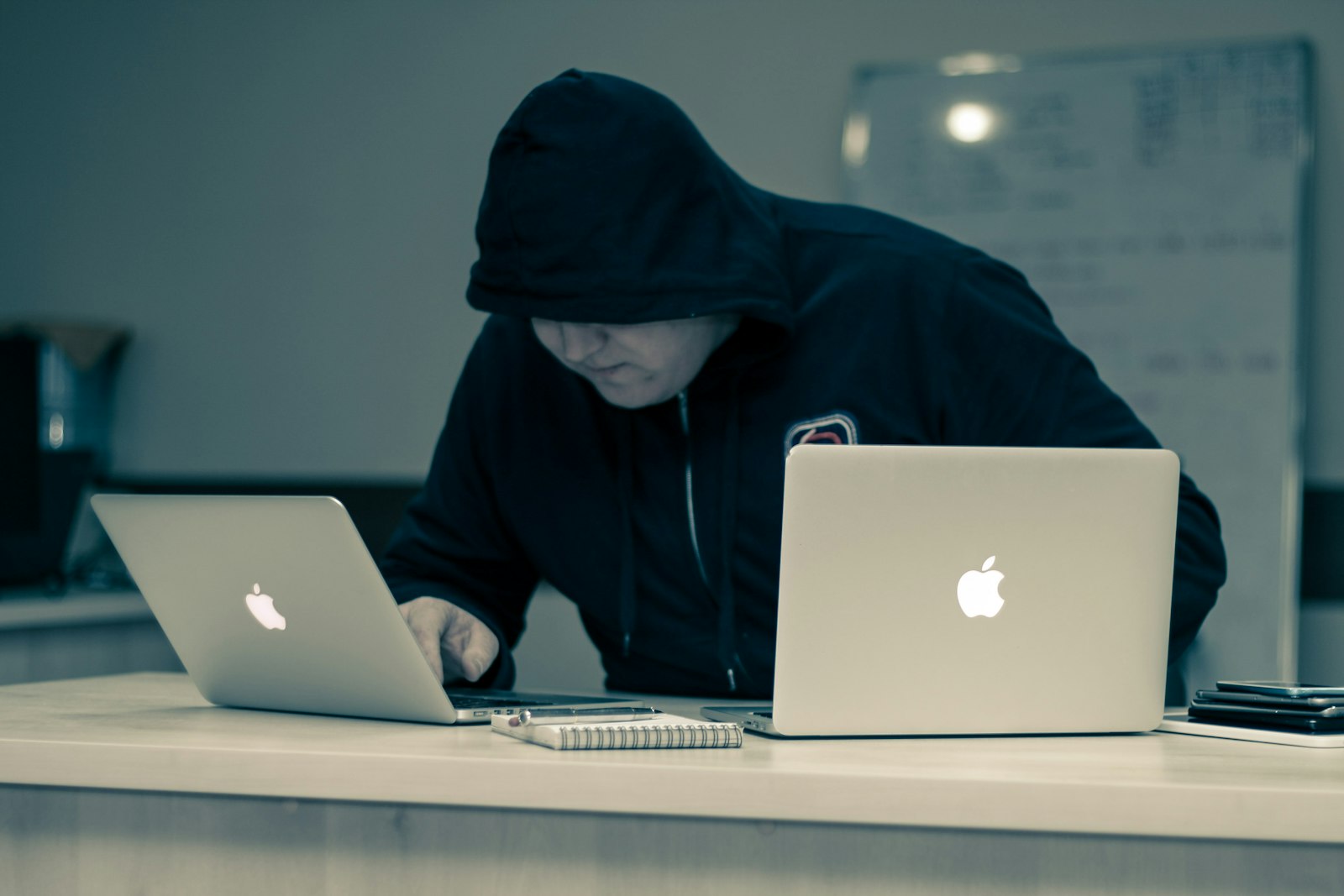 A person in a hooded sweatshirt working on two MacBook laptops in a dark room