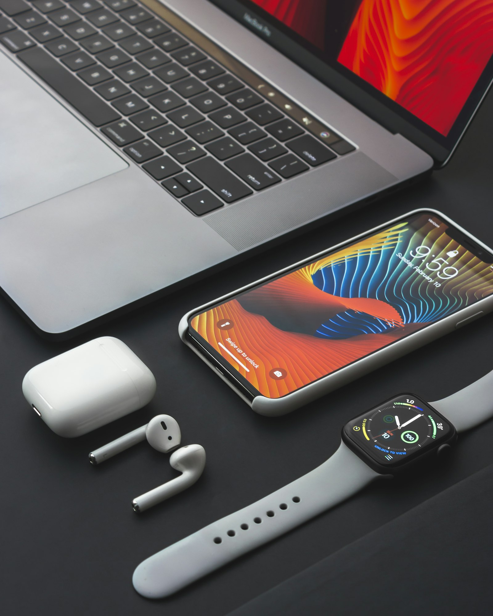 An iPhone, AirPods in their charging case and an Apple Watch arranged beside a MacBook