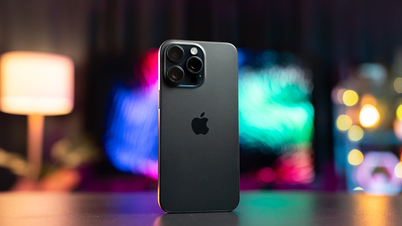 Un iPhone Pro apoyado sobre un escritorio, visto por detrás, ante luces de colores desenfocadas.