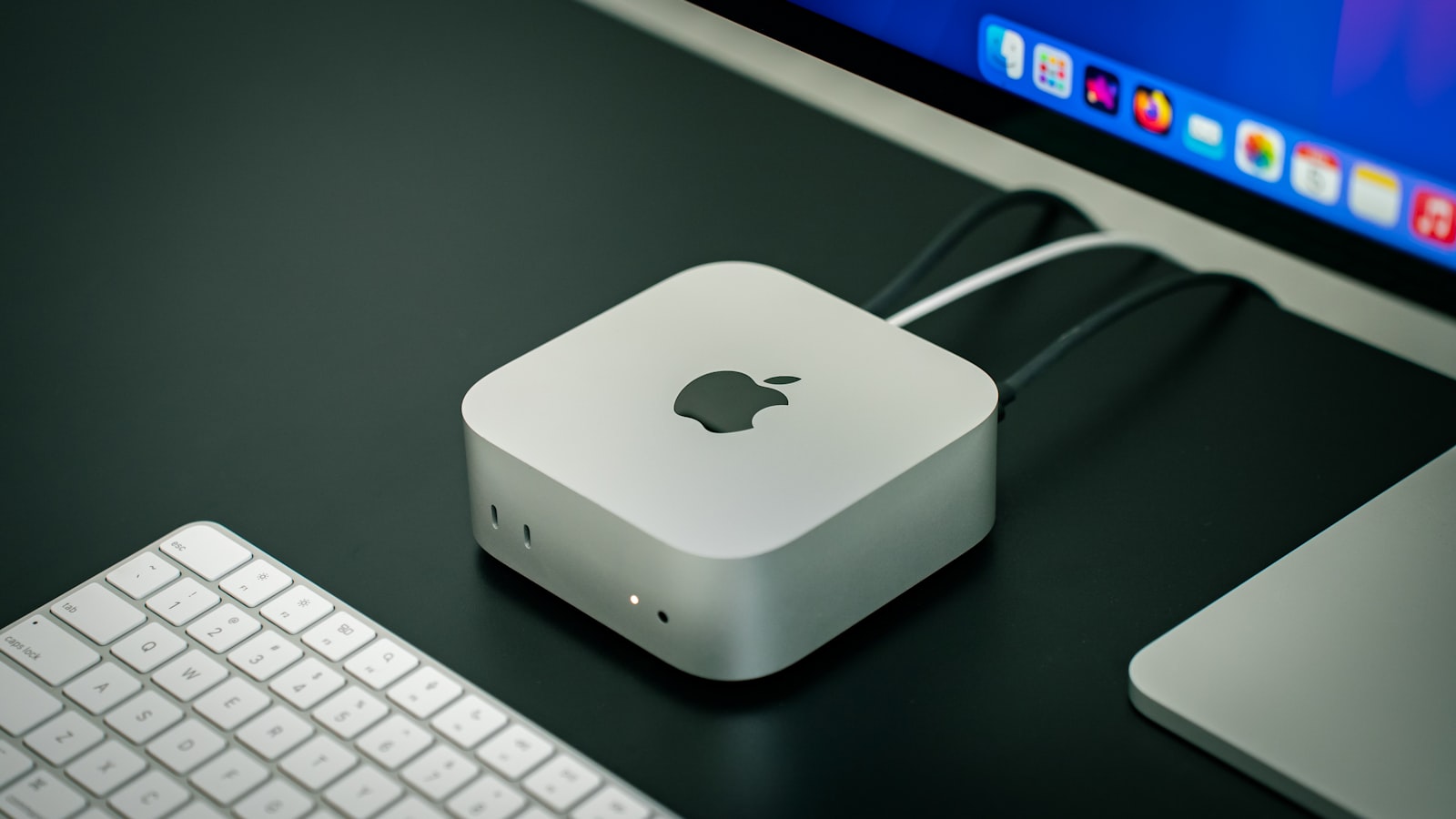 A current-generation Mac mini on a dark desk, with its front ports, keyboard and display visible