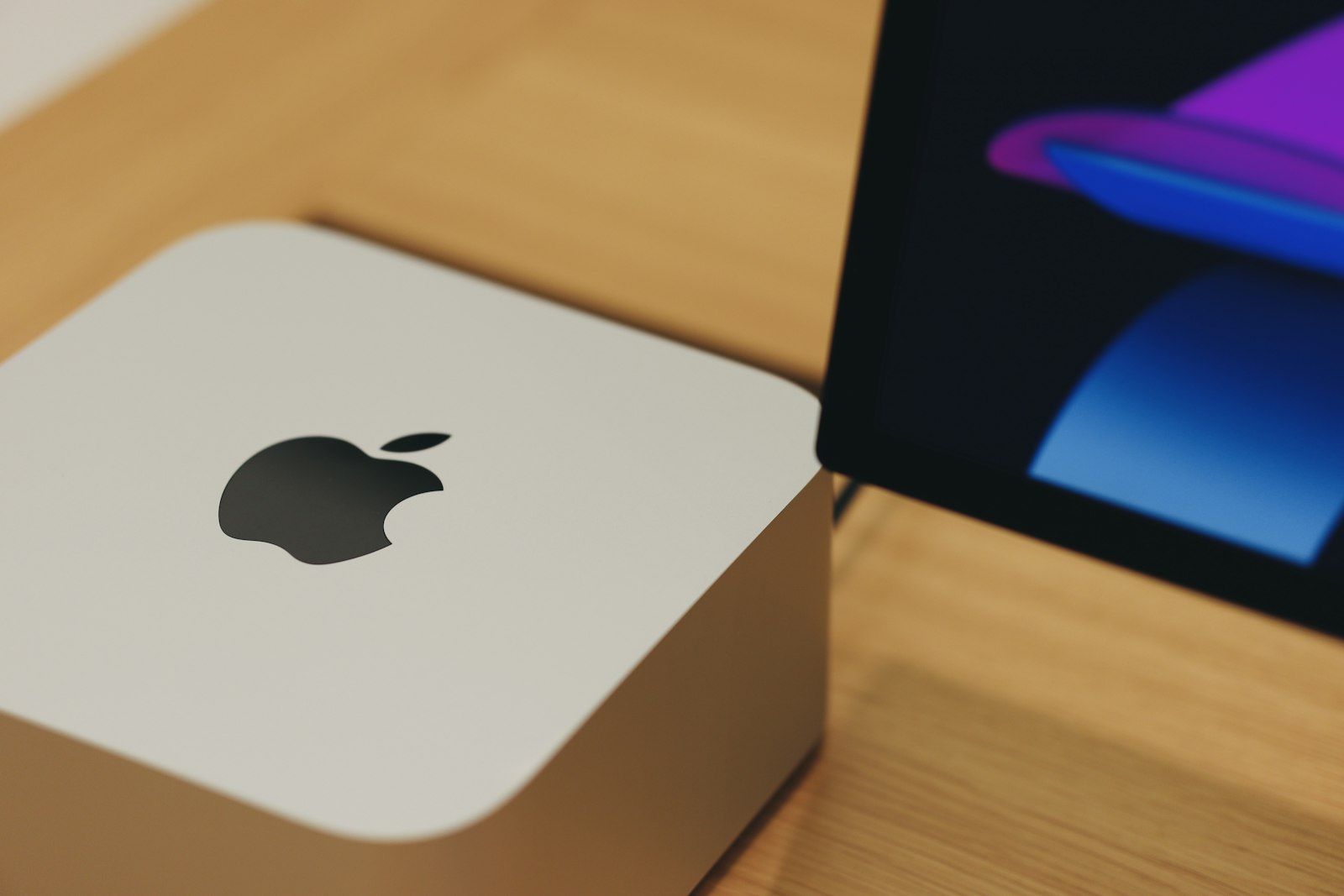 A current-generation Mac Studio on a light wooden desk, with the Apple logo visible and a display behind it