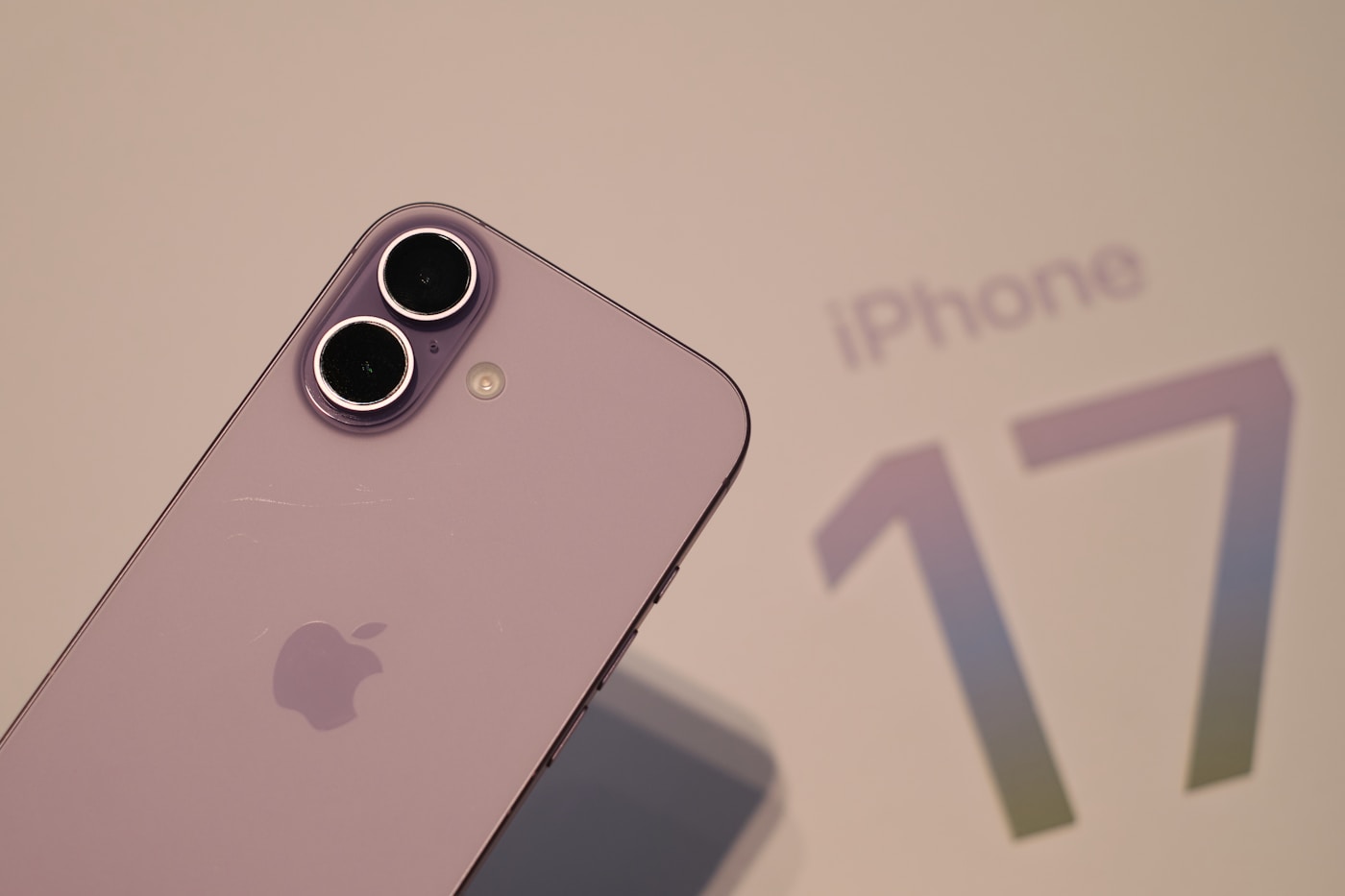 Close-up of a lavender iPhone with a vertical dual-camera module, photographed in front of an iPhone 17 display panel.