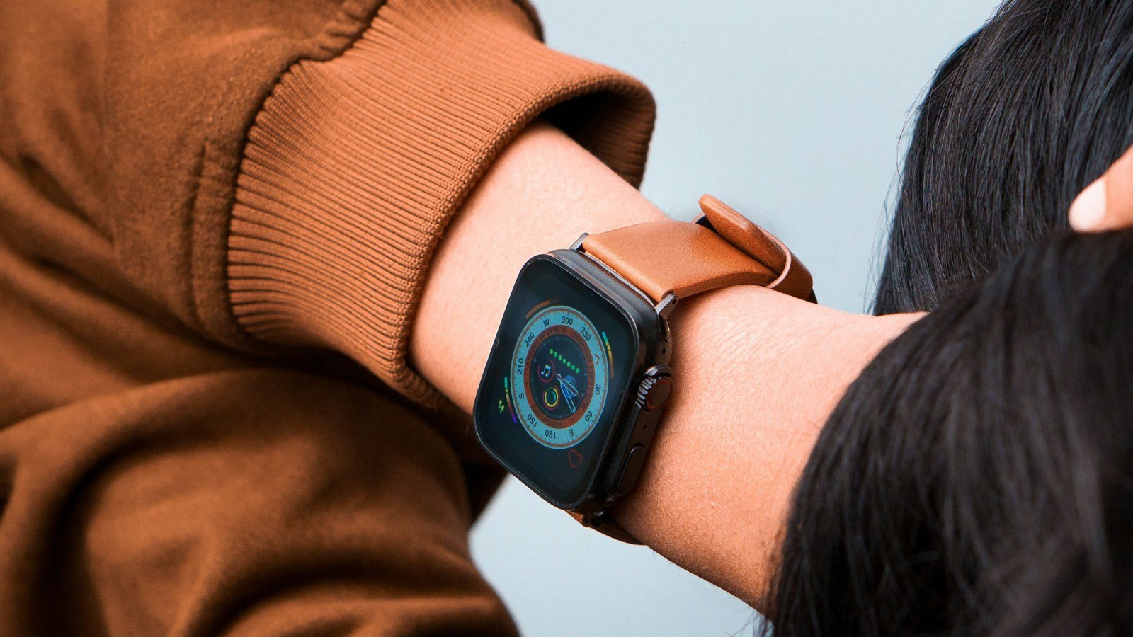 An Apple Watch with a dark case and tan leather band on a woman's raised wrist, screen lit on a watch face with activity rings; the Digital Crown and side button are visible.