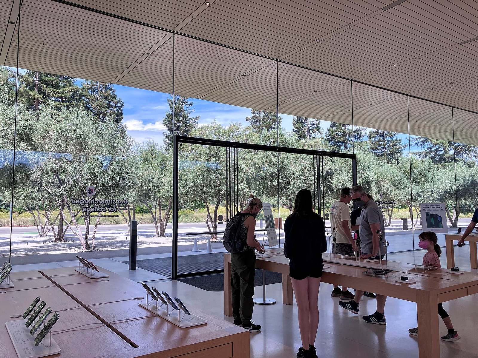 Illustration image: inside the Apple Park Visitor Center store in Cupertino, visitors at wooden tables displaying iPhones and Apple Watches, with olive trees behind the glass walls. The keynote itself was a pre-recorded video.