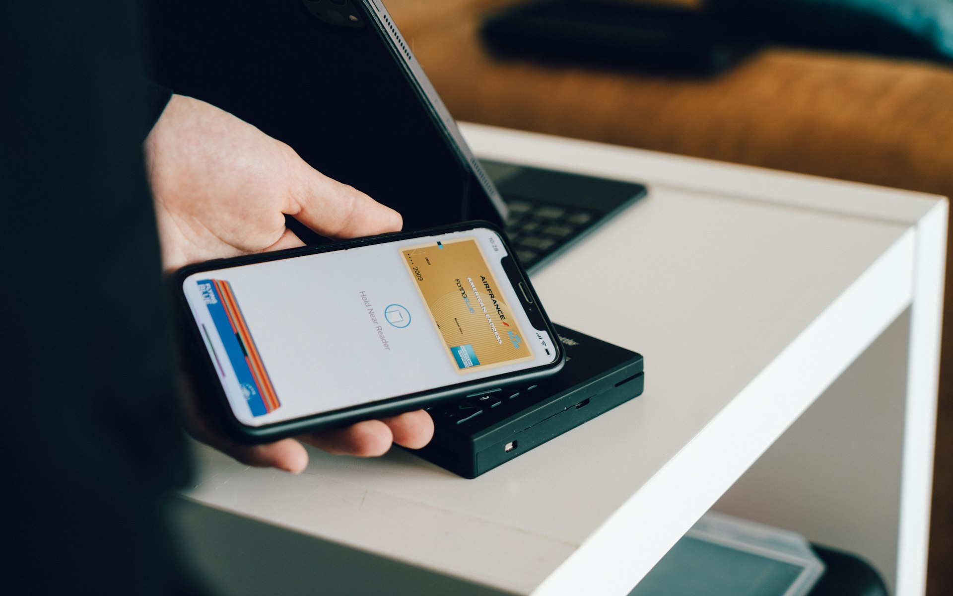 Un iPhone muestra Apple Wallet junto a un lector sin contacto; las tarjetas visibles son de pago, no un permiso de conducir.